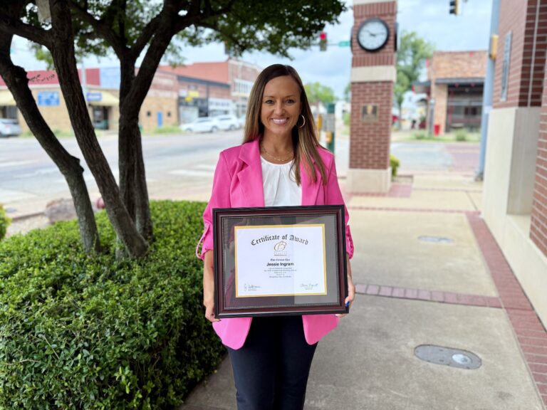 Smiling woman in a pink blazer holds a framed certificate outside on a sidewalk with greenery nearby.